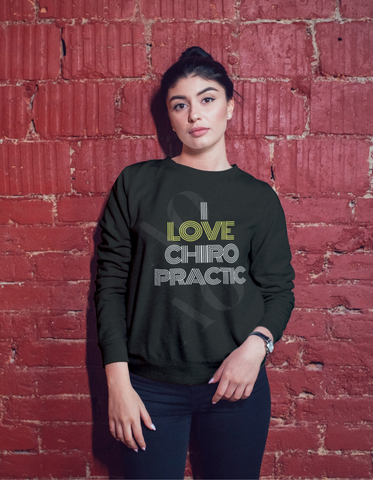 Black crewneck sweater on a woman standing against a brick wall. Crewneck says I Love Chiropractic in white letters. The word Love is in yellow letters